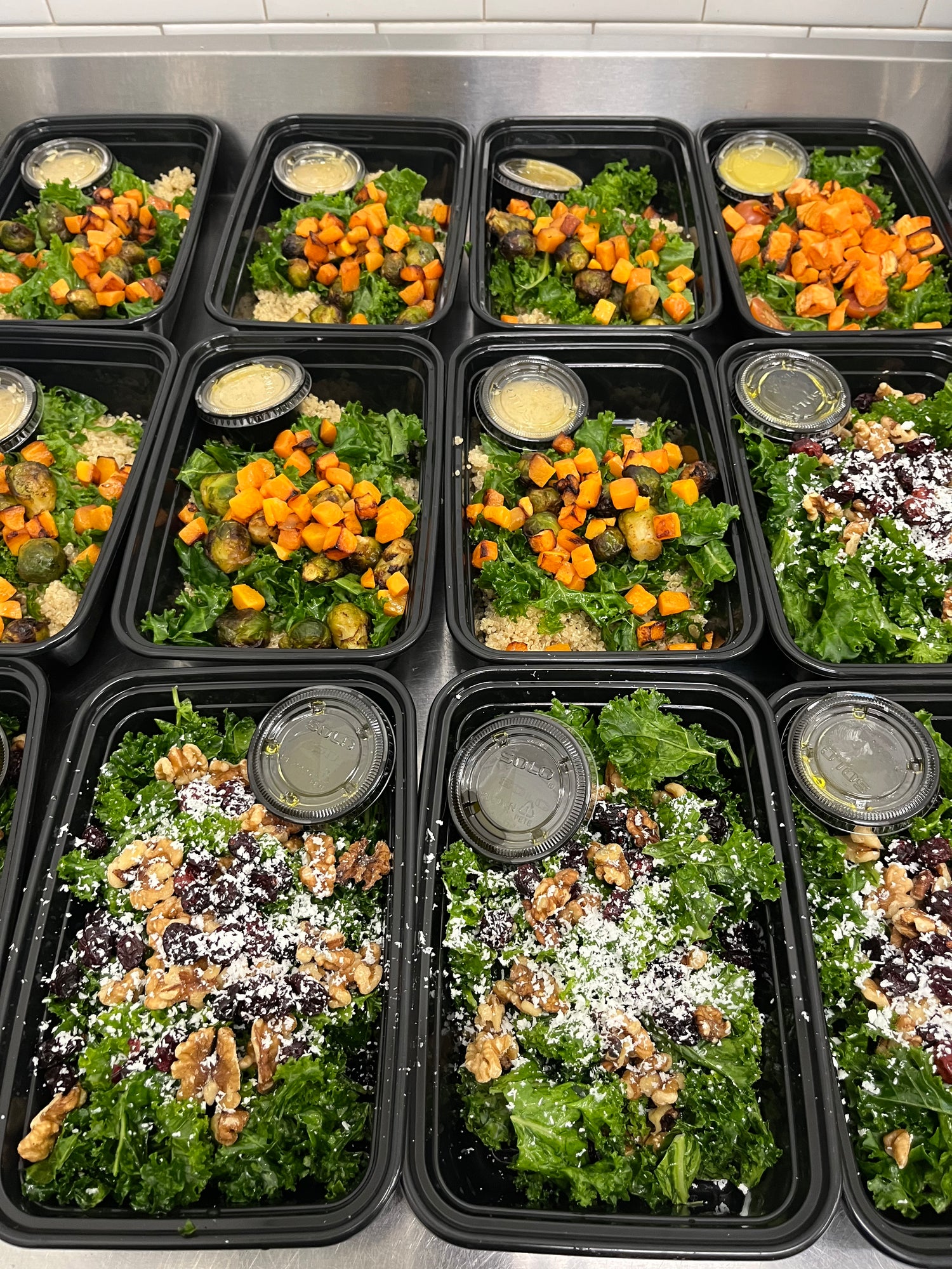 A variety of fresh salads in black containers featuring kale, roasted sweet potatoes, Brussels sprouts, walnuts, and dressings.